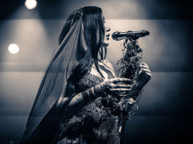Noah Cyrus performing at The Observatory in Santa Ana, vocalist in a sequined gown and veil holding a bundle of dried branches and flowers while singing into a microphone, in a black and white photo
