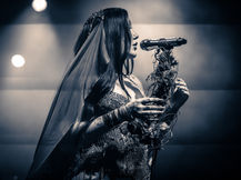 Noah Cyrus performing at The Observatory in Santa Ana, vocalist in a sequined gown and veil holding a bundle of dried branches and flowers while singing into a microphone, in a black and white photo