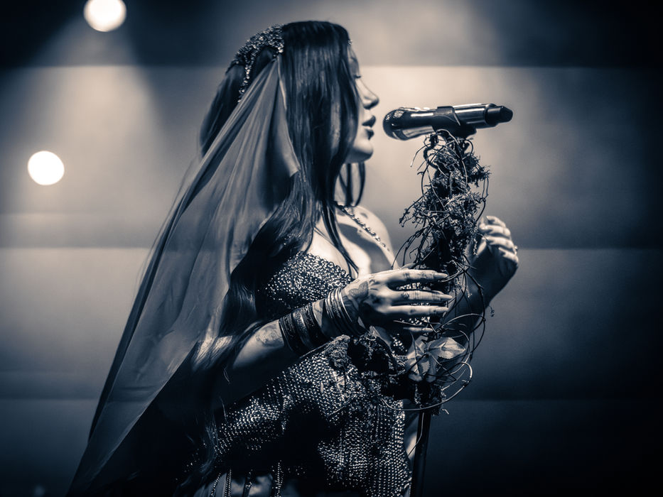 Noah Cyrus performing at The Observatory in Santa Ana, vocalist in a sequined gown and veil holding a bundle of dried branches and flowers while singing into a microphone, in a black and white photo