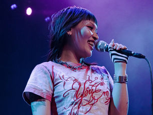 A Sobs vocalist smiling while singing into a microphone, wearing a white graphic tee with red cursive script, blue jeans with a studded belt, a silver chain necklace, large silver cuff bracelet, and zebra-print fingerless gloves, bathed in purple and blue stage lighting at Riverside Municipal Auditorium — photo by Diana Torres