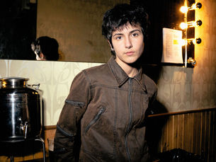 Deadbeat Girl photographed backstage in New York City, standing in a brown jacket in front of a vanity mirror with warm bulb lighting