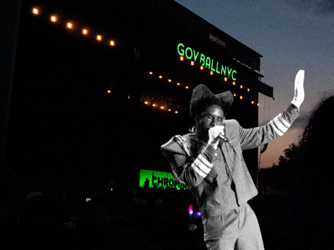 Tyler the Creator at Governors Ball at Flushing Meadows Corona Park in New York City, singing into a microphone with one arm raised in a suit and wide-brim hat at dusk, with the glowing Gov Ball NYC sign visible on the stage behind them