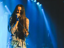Kita Alexander performing at Metro Theatre in Sydney, eyes closed and singing into a handheld microphone, long straight dark hair falling past her waist, wearing a flowing sheer lace duster over ripped black jeans, standing on a white rug on stage under dramatic blue beam lighting