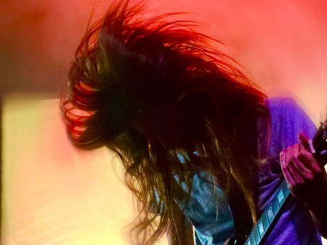Elder guitarist headbanging on stage at The Regent Theater in Los Angeles, long dark hair whipping through the air mid-motion while playing an electric guitar, wearing a purple t-shirt against swirling red and yellow stage lighting