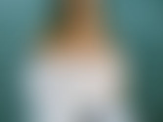 Kylee Phillips sits in front of rippling water wearing an off-the-shoulder white blouse and hoop earrings, looking directly at the camera with a steady, powerful expression, her shoulder tattoos visible in natural light.