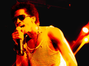 Love Spells performing at Constellation Room in Santa Ana, singer leaning into the mic with intensity, wearing a white mesh tank top, dark sunglasses, layered necklaces, and studded wristbands, bathed in deep red and orange stage lighting with dramatic crossing beams behind them, shot by Krystal Vasquez