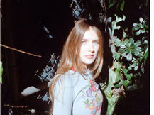 Hannah Cole standing outdoors in dappled sunlight against a chain-link fence with green foliage, wearing a light blue fitted graphic sweater with floral tattoo-style artwork and a red polka dot skirt, long straight auburn hair, looking over her shoulder at the camera, shot on film by Adam Alonzo