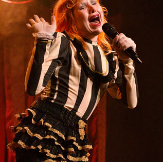 Performer in a bright orange wig and white clown-style face makeup sings with mouth wide open and one hand raised, holding a microphone in a white fishnet glove. They wear a black-and-cream striped top and black ruffled shorts with gold lace trim on stage at Varsity Theater in Minneapolis. Photo by Kailey Blunk.