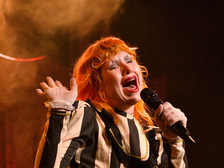 Haute & Freddy performing at Varsity Theater in Minneapolis, singing intensely into a handheld microphone with eyes closed and one hand raised, wearing a black and white vertical striped long-sleeve top, tiered black and gold ruffle skirt, black tights, and mesh lace gloves, with warm amber stage lighting and a red curtain in the background