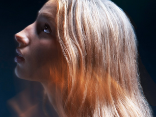 Ali Wolverton in profile against a deep blue-black background, golden blonde hair catching warm light as she gazes upward in a moody, cinematic portrait.