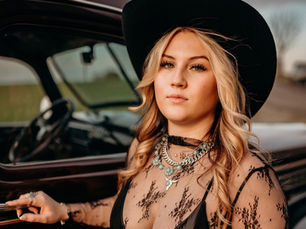 Lilly Moss leaning against a vintage dark car in a black lace sheer top, wide-brim cowboy hat, and layered turquoise and chain necklaces in a moody, golden-hour outdoor portrait.