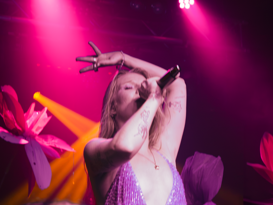 Lilyisthatyou performing on stage at the Oslo in Hackney, London, wearing a silver sequin deep-V dress with arms raised overhead, tattoos visible on both arms, singing into a microphone surrounded by oversized pink and red floral stage props under hot pink and orange lighting