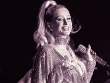 Paris Hilton performing at Outloud Festival at West Hollywood Park in Los Angeles, smiling while holding a microphone in a sequined outfit with a rhinestone choker and high ponytail, in a purple-toned photo