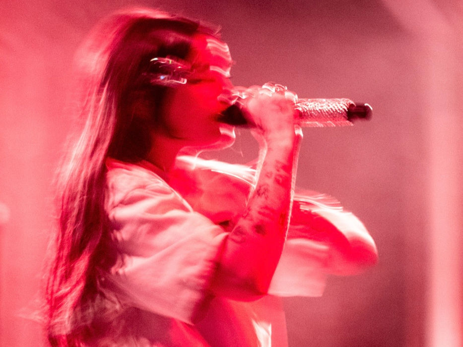 BENEE performing on stage at the Varsity Theater in Minneapolis, wearing a white ruffled tiered dress and singing into a bedazzled microphone, captured in intentional motion blur under deep red stage lighting