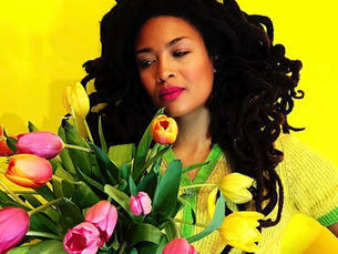 Valerie June holding a large bouquet of pink, yellow, and orange tulips against a bright yellow background, wearing a yellow and green knit top, voluminous locs framing her face, eyes softly downcast with a slight smile and pink lips