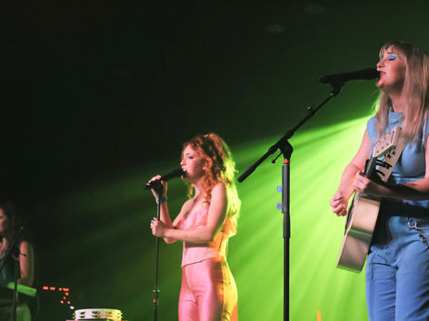 Trousdale performing at The Senate in Columbia SC, three women on stage — one on keys, one singing into a microphone in a pink outfit, one playing acoustic guitar and singing — under green stage lighting