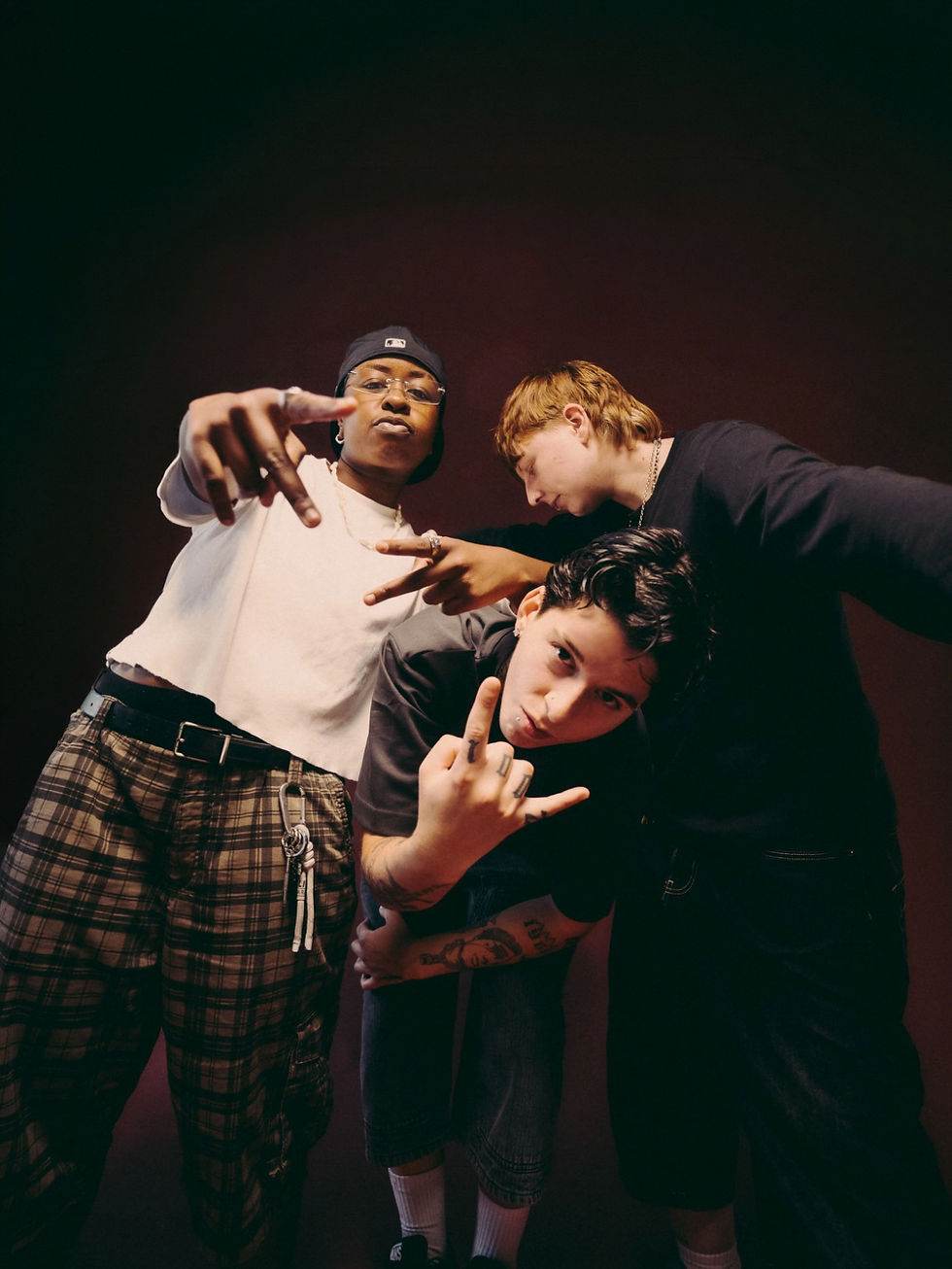 Three members of UNINVITED pose close together against a dark red backdrop, making hand signs toward the camera. On the left, one wears a backward navy cap, clear glasses, a white cropped tee, and plaid cargo pants with a carabiner of keys. In the middle, one with dark swept hair and tattooed arms leans forward, and behind them one with tousled auburn hair in a black long-sleeve leans in with a peace sign.
