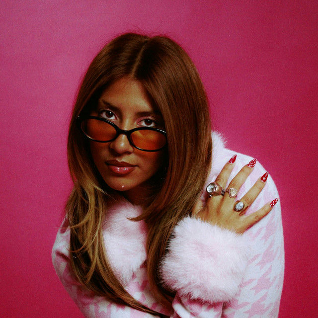 Mia Lailani posing against a bright pink background, wearing narrow orange-tinted sunglasses and a pink fuzzy houndstooth sweater, hand resting on her shoulder showing multiple silver rings and long red stiletto nails with polka dot detail, long brown hair falling past her shoulders