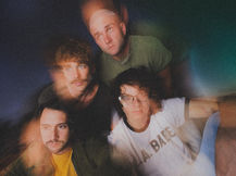 Promo photo of The Wrecks: four band members crowded close together looking toward the camera, with soft blur and prismatic blue, green, and orange light leaks over a dark background. One wears a yellow tee and olive jacket, another a white "L.A. BABE" tee and glasses.