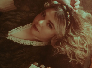 Lounna lying down holding the neck of a white guitar, wearing a dark lace dress and a floral headband, looking up at the camera in a warm, golden-toned intimate portrait.