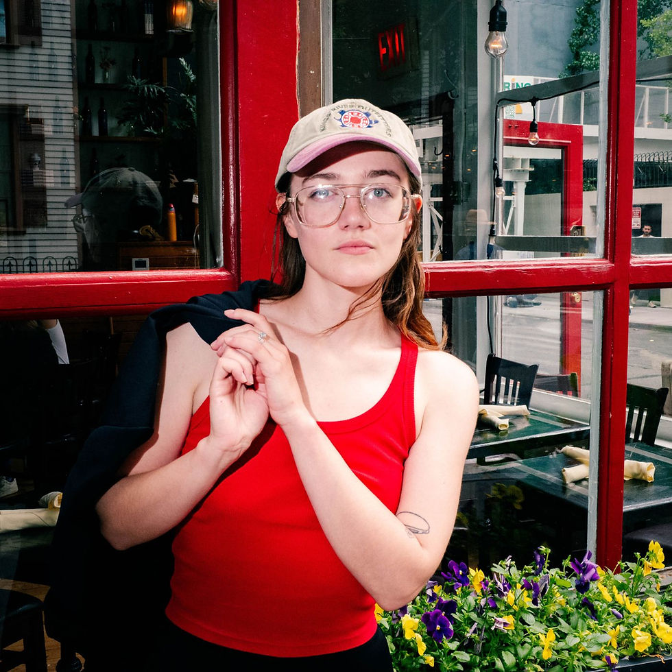 Moselle stands outside a café with red-framed windows, wearing a red tank top, glasses, and a baseball cap, holding a jacket over her shoulder while looking calmly at the camera, with flowers in the foreground.