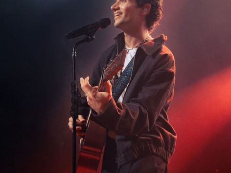 Jeremy Zucker performing at Brooklyn Steel in New York City, smiling at a microphone stand while holding an acoustic guitar in a brown zip-up jacket under red and blue stage lighting