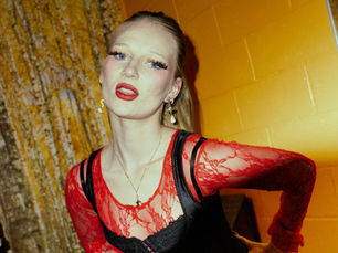 Lilyisthatyou photographed backstage at Elsewhere in Brooklyn, wearing a red lace top and black dress with dramatic makeup and Vivienne Westwood earrings against a warm orange wall