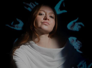 Audrey Powne seated in a chair and lit by a tight spotlight against a black background, wearing a white long-sleeve off-shoulder top with legs crossed, with blue-tinted shadowy figures projected behind her head