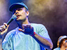 Mac DeMarco performing at The Greek Theater in Los Angeles, vocalist in a grey t-shirt and baseball cap singing into a microphone with hand outstretched, guitarist visible behind him under blue and amber stage lighting