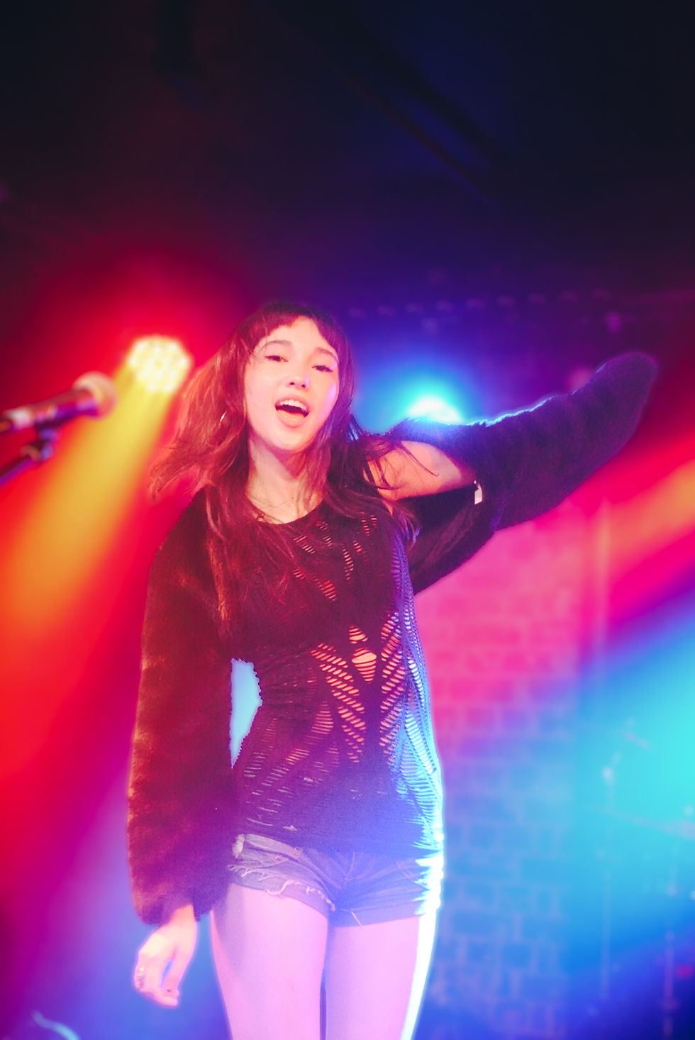 Jade Yee Smith performing at Lansdowne Hotel in Sydney, singing with her arm raised and head tilted back, long wavy reddish-brown hair with bangs, wearing a black distressed cut-out knit top with fuzzy long sleeves and denim shorts, surrounded by vibrant red, blue, and purple stage lighting