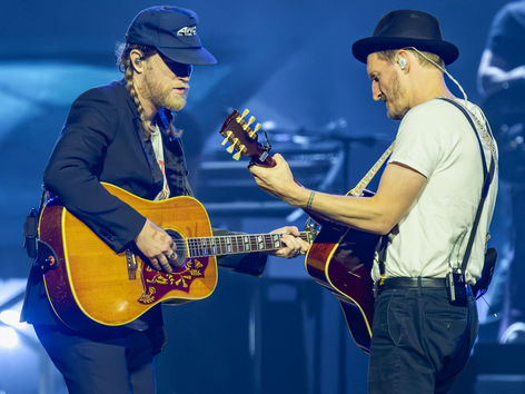 The Lumineers performing at Kia Forum in Los Angeles, two guitarists facing each other on stage playing acoustic guitars under blue stage lighting