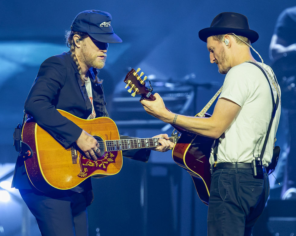 The Lumineers performing at Kia Forum in Los Angeles, two guitarists facing each other on stage playing acoustic guitars under blue stage lighting