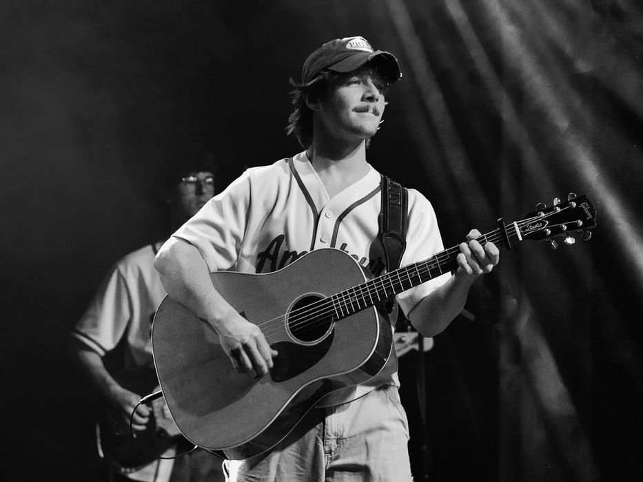 Brenn! playing an acoustic guitar on stage at the Varsity Theater in Minneapolis, wearing a baseball jersey and cap with a thin mustache, looking upward mid-performance, in black and white with a second guitarist visible in the background