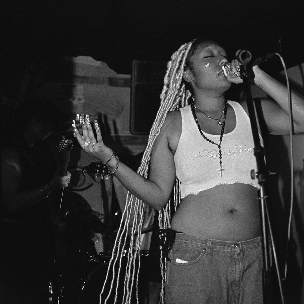 Eph See performs on stage in a black-and-white photo, singing into a microphone with eyes closed, wearing a cropped tank top and layered necklaces, with long braided hair and a band playing in the background.