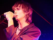 Park Rd vocalist singing into a handheld microphone on stage at Lansdowne Hotel in Sydney, medium-length shaggy brown hair, wearing a dark pinstripe blazer open over a bare chest with a delicate black drop necklace and earrings, bathed in warm pink and red stage light with teal blue beams behind them, guitar neck visible at the edge of the frame