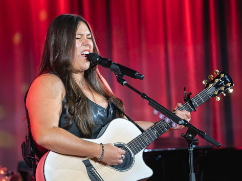 Gigi Perez performing at the Grammy Museum in Los Angeles, singing with eyes closed into a microphone while playing a white acoustic guitar against a red curtain backdrop