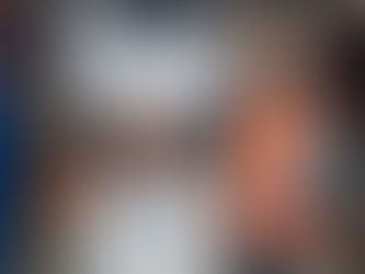 Worker in orange vest and hard hat uses a tablet in a warehouse. Two others in background with boxes. Bright lighting, organized shelves.