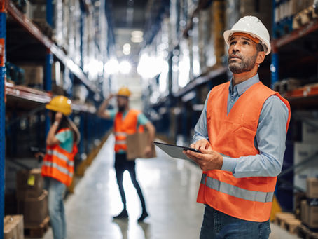Worker in orange vest and hard hat uses a tablet in a warehouse. Two others in background with boxes. Bright lighting, organized shelves.