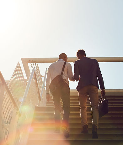 Two business professionals walking up a staircase together in bright sunlight, seen from behind — one in a blazer holding a briefcase, the other in casual businesswear with a messenger bag. The scene conveys partnership, mentorship, and forward momentum, reflecting KLV Accounting’s supportive client relationships and business advisory services in Calgary, Alberta.