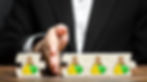 A hand separates puzzle pieces with money icons on a wooden table. A person in a black suit is in the background, suggesting financial decisions.
