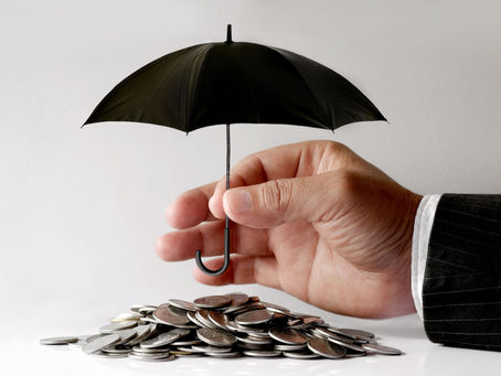 A hand holds a small black umbrella over a pile of coins, suggesting financial protection. The background is plain, emphasizing the objects.