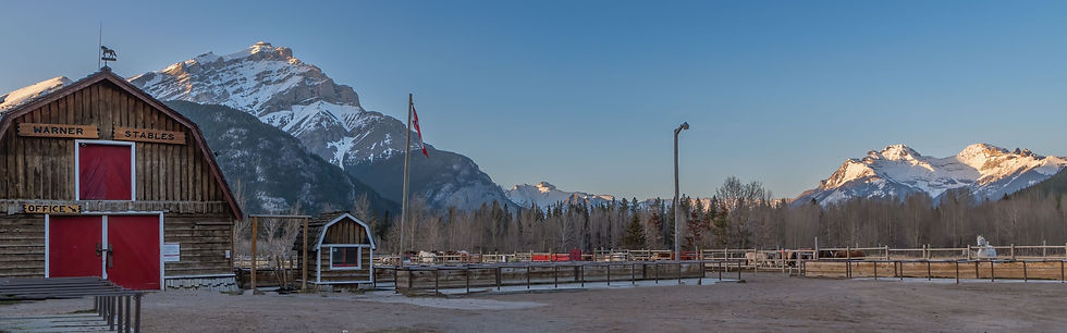 Rustic barn marked “Warner Stables” with red doors and a Canadian flag, set against a backdrop of snow-covered Rocky Mountains and clear blue skies. Surrounded by fences and horses, the scene captures Alberta’s rural charm and western heritage — reflecting the local roots and small business focus supported by KLV Accounting in Calgary.