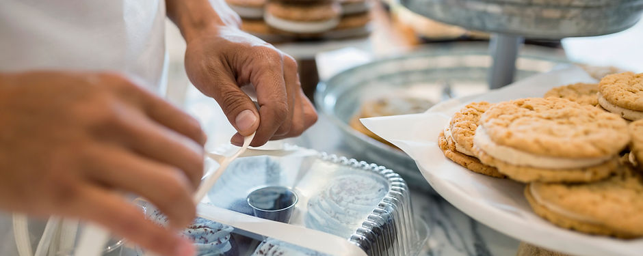 A person's hands are shown tying a white ribbon around a clear plastic container of chocolate cupcakes with white frosting. In the background are several oatmeal cookies and other pastries on a table.