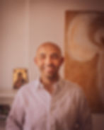 Ilias Moschos lächelt im weißen Hemd vor einer hellen Wand mit einem Kunstwerk im Hintergrund