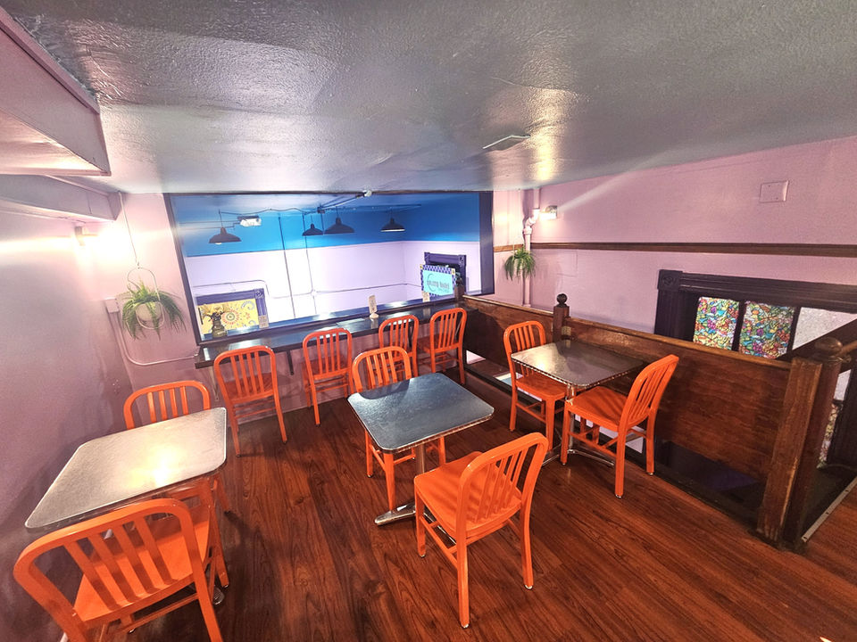 Three tables with two orange chairs each sit in front of a counter with several more chairs. 