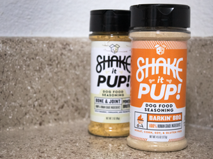 This image features two containers of "Shake it Pup!" dog food seasoning placed on a beige speckled countertop against a white wall. The container in the foreground is labeled "Barkin' BBQ" with an orange and white design, highlighting that it contains 100% human-grade ingredients and is free from wheat, corn, soy, and gluten. The second container in the background has a yellow and white design and is labeled "Bone & Joint," likely indicating a health-focused variety. Both containers have black flip-top lids