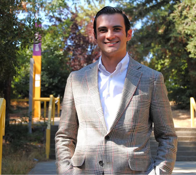 Brandon Brookshire wearing a gray suit and white shirt standing on a stairway with several trees in the background.