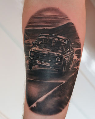 A black and grey tattoo of a rally car on an arm. Made in Llangefni, Isle of Anglesey, North Wales by Shelley Williams.