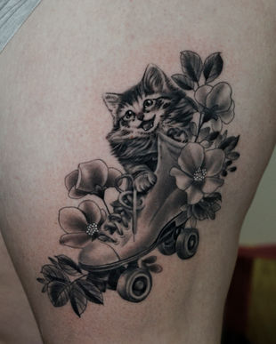 A black and grey tattoo of a cheeky kitten in a roller skate with flowers on a leg, made from the clients own drawn reference. Made in Llangefni, Isle of Anglesey, North Wales by Shelley Williams. 