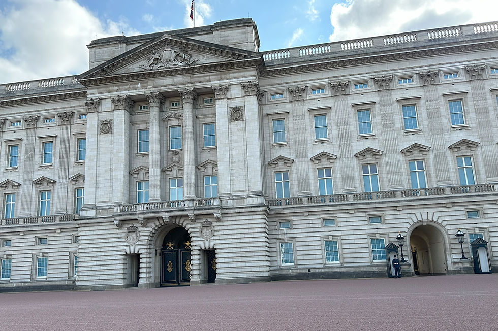 Buckingham Palace facade under a partly cloudy sky, with a guard standing at attention near an arched entrance.